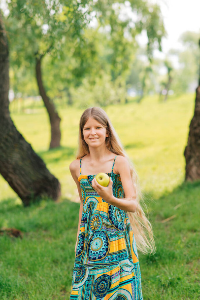 cute young girl with beautiful long hair looks at an apple in her hand.  Summer day. The child collects fruits in the garden. Fresh healthy food for children. Family meals in the summer. - Фото, зображення