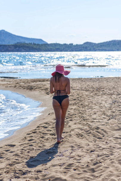 задний вид сексуальной женщины в черном бикини и красной шляпе, идущей по песку тропического пляжа - Фото, изображение
