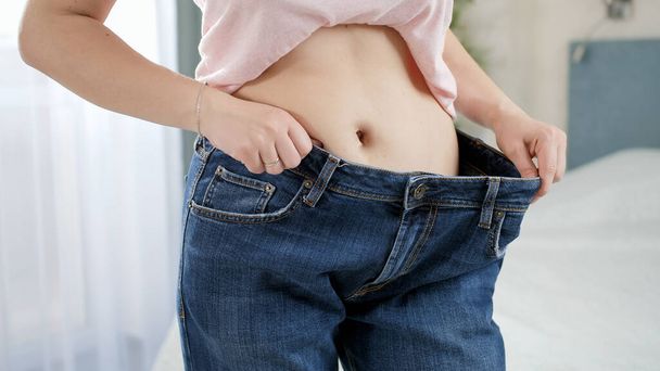 Beautiful young woman with flat stomach posing in front of mirror wearing old large jeans. Concept of dieting, loosing weight and healthy lifestyle. - Foto, afbeelding