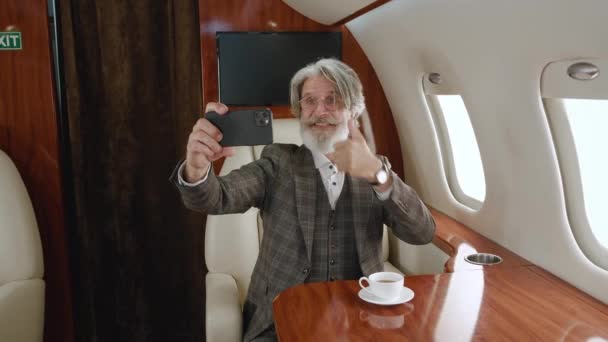 Homem rico maduro usando smartphone para chamadas de vídeo durante viagens a jato privado de luxo. Homem sênior de cabelos grisalhos elegante ter videoconferência no avião - Filmagem, Vídeo