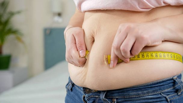 Jonge vrouw lijdt aan overgewicht meten van haar dikke buik. Concept van dieet, ongezonde levensstijl, overgewicht en obesitas - Foto, afbeelding