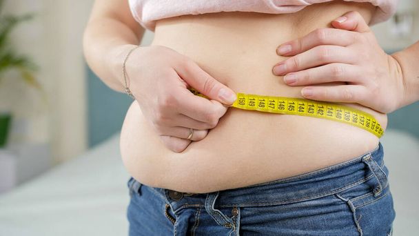 Zbliżenie kobiety z nadwagą przy użyciu taśmy pomiarowej. Pojęcie diety, niezdrowego trybu życia, nadwagi i otyłości - Zdjęcie, obraz