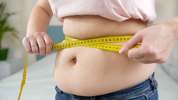 Primer plano de la mujer obesa midiendo su gran vientre con cinta métrica amarilla. Concepto de dieta, estilo de vida poco saludable, sobrepeso y obesidad - Foto, Imagen