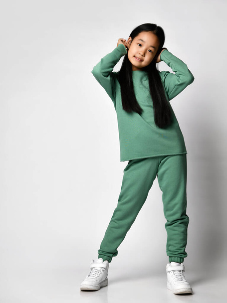 Children, lifestyle and body language. Full length asian girl with long hair in a comfortable casual green suit. Baby girl stands on a light background and straightens her hair - Zdjęcie, obraz