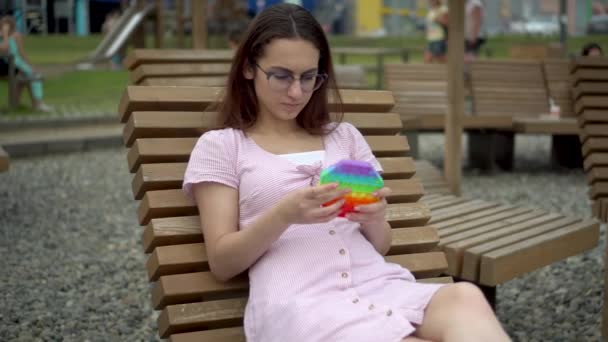 Angry young woman in glasses lies on a deck chair with pop it in her hands. The girl plays popit to calm down. - Footage, Video