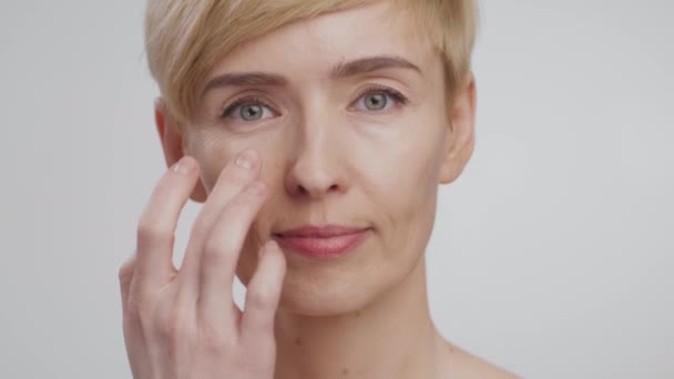 Close up portrait of beautiful middle aged woman applying anti-wrinkle cream on under eyes area, looking at camera - Footage, Video