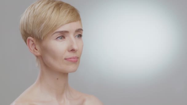 Semi profile portrait of happy middle aged blonde lady looking aside at empty space and smiling, grey background, mockup - Footage, Video
