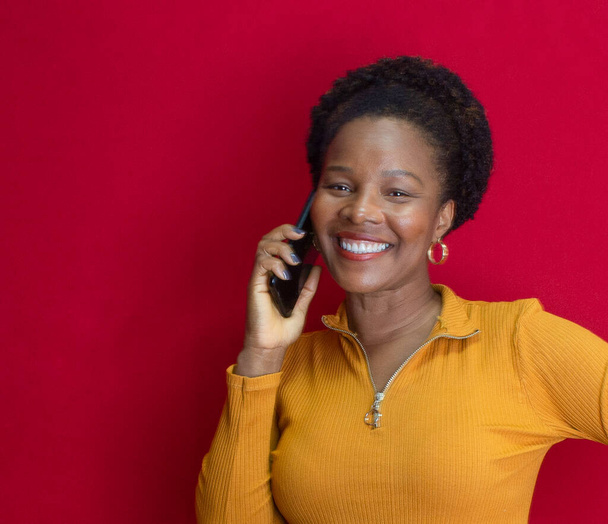 schwarze Frau lächelt und hält ein Handy mit gelbem Hemd vor rotem Hintergrund - Foto, Bild