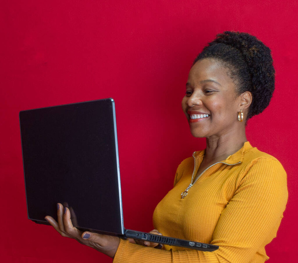 černá žena s úsměvem drží na notebooku se žlutou košilí v červeném pozadí - Fotografie, Obrázek