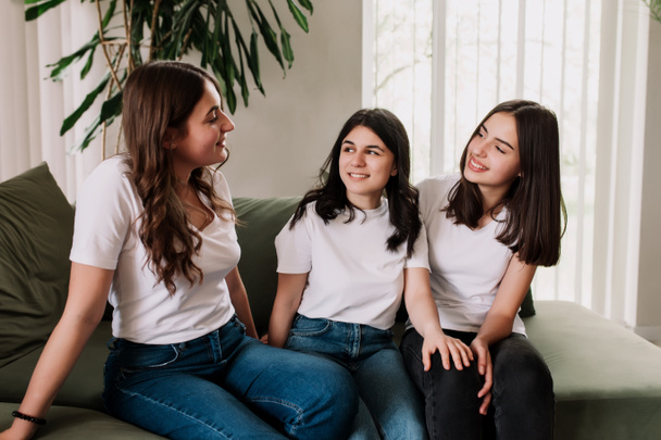 Three pretty smiling caucasian girls in white shirt sitting on the green sofa. Friends emotionally talking and discussing last news and life, sharing their thoughts and having some gossip topics - Photo, Image