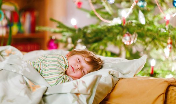 Mały słodki blondyn śpiący pod choinką i marzący o Mikołaju w domu, w domu. Tradycyjny festiwal chrześcijański. Szczęśliwy dzieciak czeka na prezenty. - Zdjęcie, obraz