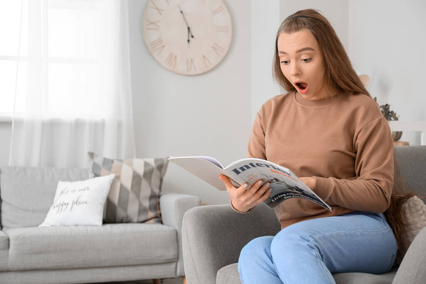 Σοκαρισμένη νεαρή γυναίκα διαβάζει περιοδικό στο σπίτι - Φωτογραφία, εικόνα