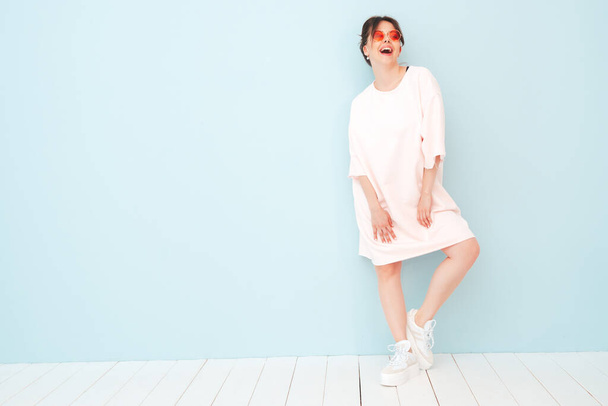 Portrait of young beautiful smiling female in trendy summer pink clothes. Sexy carefree woman posing near light blue wall in studio. Positive model having fun indoors - 写真・画像