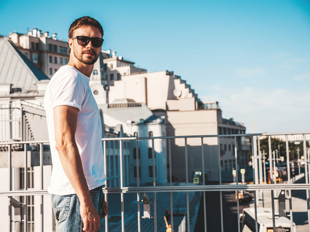 Portrait of handsome confident hipster lambersexual model.Stylish man dressed in white T-shirt. Fashion male posing behind blue sky on the street background in sunglasses - Foto, immagini