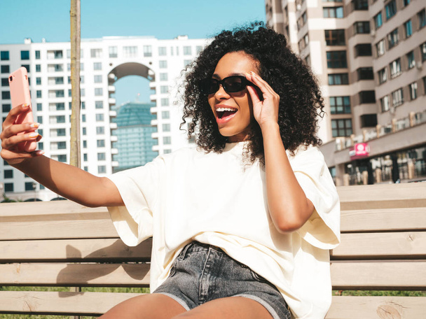 Beautiful black woman with afro curls hairstyle.Smiling hipster model in white t-shirt. Sexy carefree female posing in the street in sunglasses. Cheerful and happy.Taking selfie photo - Zdjęcie, obraz