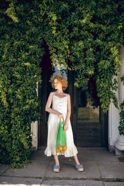 Young woman in a white long dress holding a green mesh bag in her hands - Foto, afbeelding