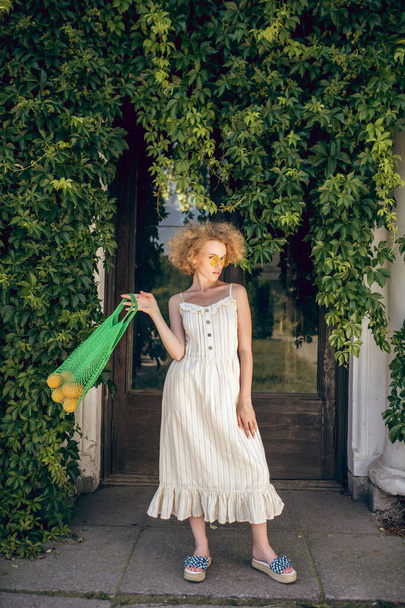 Young woman in a white long dress holding a green mesh bag in her hands - Foto, afbeelding