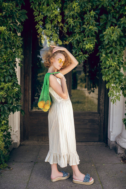 Pretty woman in a white long dress and sunglasses holding a green mesh bag in her hands - Foto, Imagen