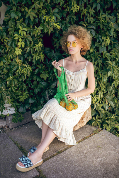 Pretty woman in a white long dress and sunglasses holding a green mesh bag in her hands - Fotoğraf, Görsel
