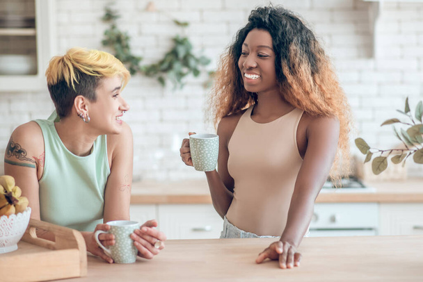 Two cheerful girlfriends talking in kitchen with coffee - Foto, afbeelding
