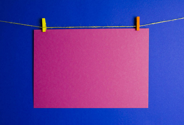 une feuille de papier rouge est suspendue sur une corde sur un fond bleu. le carton est maintenu sur une broche jaune et orange - Photo, image