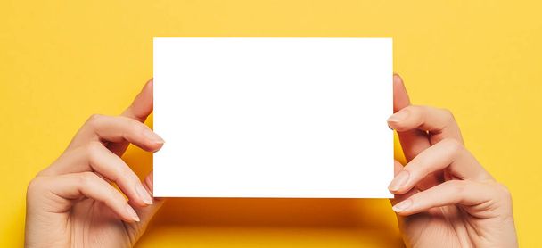 Le mani femminili tengono un foglio di carta bianco su uno sfondo giallo. Spazio pubblicitario - Foto, immagini