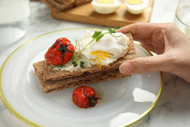 Woman eating fresh rye crispbread with poached egg, cream cheese and grilled tomato at table, closeup - Foto, immagini