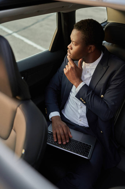 Pensive μαύρο υπάλληλο που εργάζονται σε φορητό υπολογιστή στο εσωτερικό του αυτοκινήτου - Φωτογραφία, εικόνα