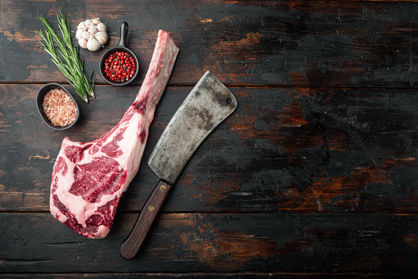 Syrový tomahawk hovězí steak se ingrediencemi na grilování se sadou na tření koření a starý řeznický sekáček, s koření a bylinkami, na starém tmavém dřevěném stolku, horní pohled plochý ležel, s kopírovacím prostorem pro text - Fotografie, Obrázek