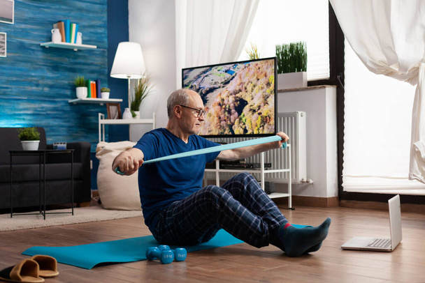 Пенсионер работает на сопротивление тела тренируя мышцы рук используя упругую ленту сидя на циновке yoga - Фото, изображение