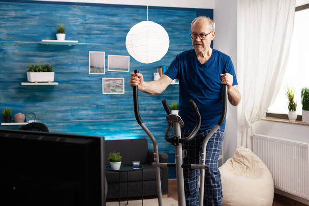 Aktív időskorú férfi edzés lábak izomellenállás kerékpár gép a nappaliban - Fotó, kép