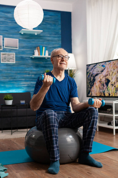 Retirement senior man sitting on swiss ball exercising arms muscles doing fitness exercises - Photo, Image