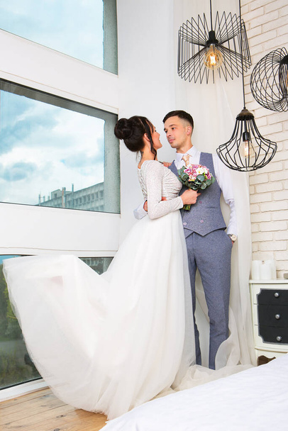 Gyönyörű menyasszony és vőlegény ölelés és csók egymást a szobában, közel az ablakhoz. - Fotó, kép