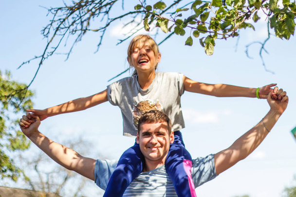 Defocused brother riding sister on back. Portrait of happy girl on man shoulders, piggyback. Girl fly, raise hands. Family playing outdoor. Green nature background. Out of focus - Photo, Image