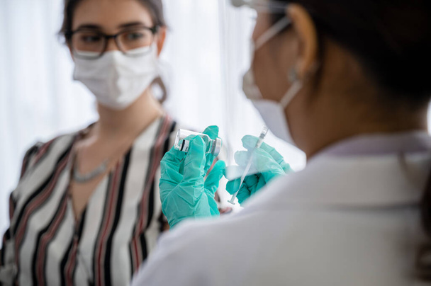 Жінка-лікар або медсестра, яка носить маску, рукавички та козирок, тримає вакцину проти коронавірусу 19 років, а жінка сидить у лабораторії в очікуванні вакцини від ін'єкцій. Концепція запобігання COVID-19
. - Фото, зображення