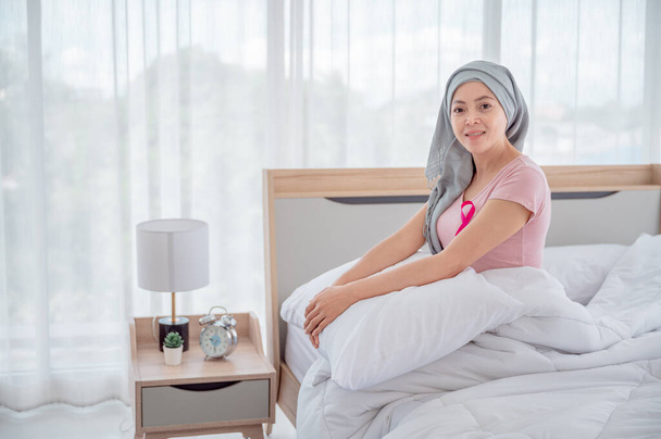 Ασιάτισσα κοπέλα που φοράει μαντίλα κάθεται στο κρεβάτι κοιτάζοντας έξω από το παράθυρο Δείχνει ανάκαμψη από καρκίνο του μαστού έννοια του καρκίνου του μαστού έννοια της πρόληψης του καρκίνου - Φωτογραφία, εικόνα