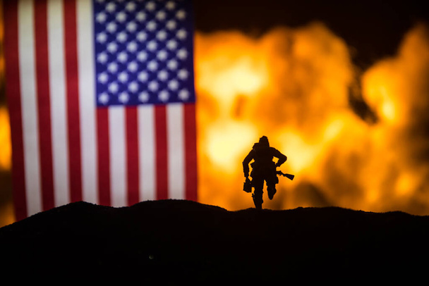 US small flag on burning dark background. Concept of crisis of war and political conflicts between nations. Silhouette of armed soldier against a USA flag. Selective focus - Photo, image
