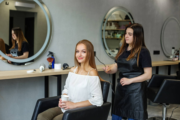 A young woman visited a hairdresser. The woman is drinking coffee and smiling. Hair styling and relaxation. - Photo, Image