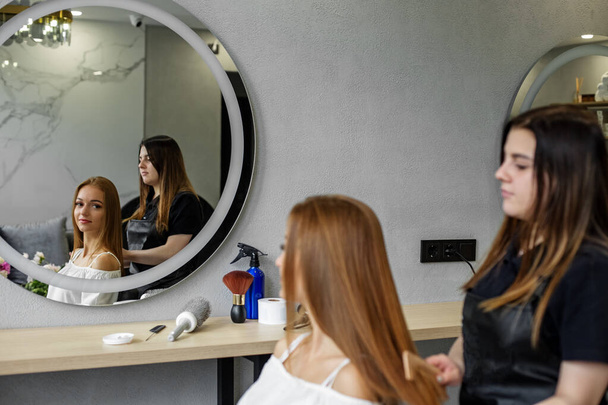 A young woman visited a hairdresser. The girl looks in the mirror. Hairdresser makes hair styling. - Photo, Image