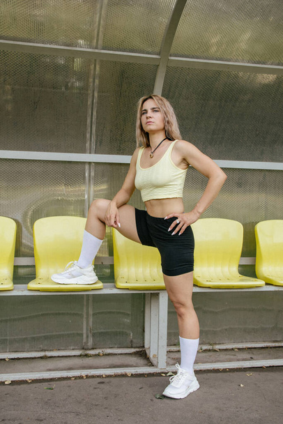 Blonde girl in sports uniform sits on yellow seats for fans - 写真・画像