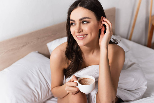 brunette woman smiling while adjusting hair and holding cup of coffee in bed - Fotoğraf, Görsel