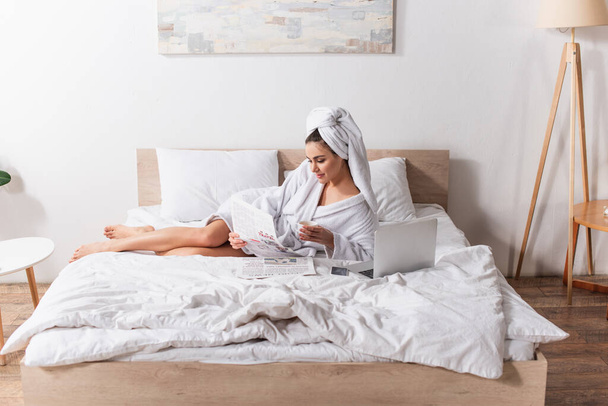 jeune femme en peignoir et serviette sur la tête tenant tasse de café et journal près de gadgets sur le lit  - Photo, image