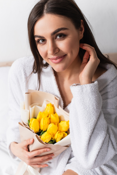joyful woman in bathrobe holding bouquet of yellow tulips in bedroom - Фото, изображение