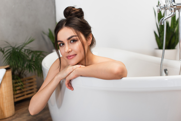 cheerful young woman with hair bun looking at camera in bathtub  - Foto, Imagem