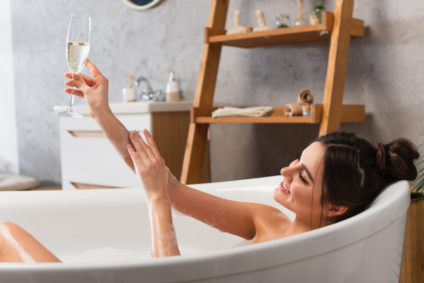 happy woman holding glass of champagne while taking bath in bathtub - Fotó, kép
