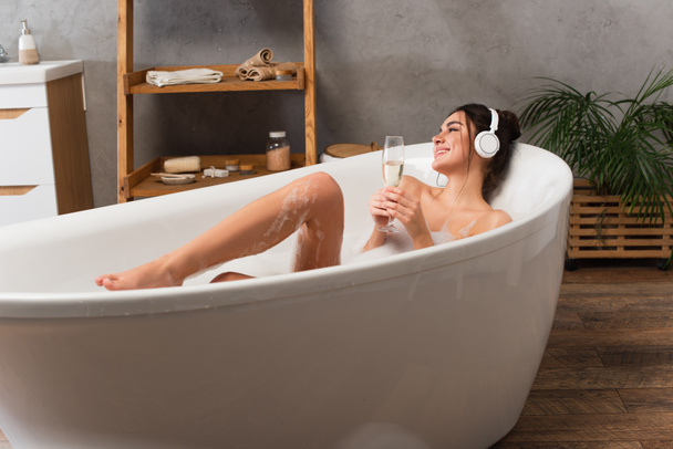 pleased woman in headphones listening music and holding glass of champagne while taking bath  - Fotó, kép