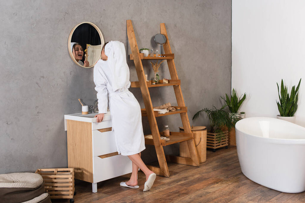mulher em roupão de banho com toalha na cabeça escovando os dentes no banheiro perto do espelho - Foto, Imagem