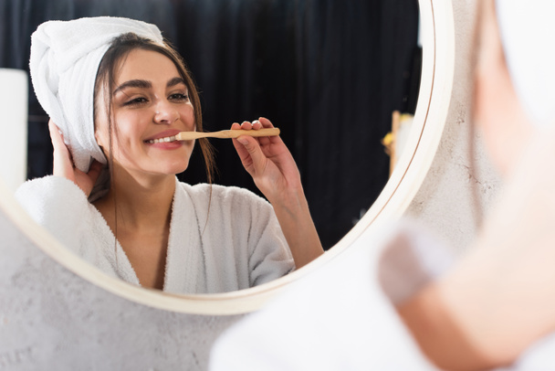 reflection of happy woman in bathrobe with towel on head brushing teeth near bathroom mirror - Foto, Bild