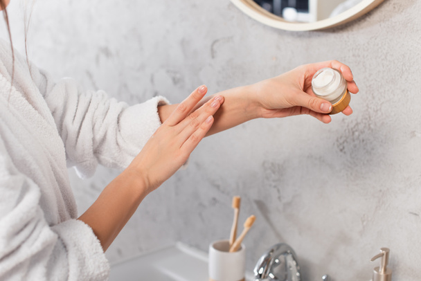 cropped view of young woman in bathrobe holding container and applying cosmetic cream  - Foto, afbeelding