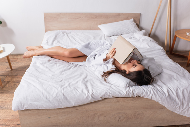 happy woman in bathrobe lying on bed and reading book - Photo, Image